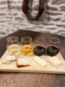 4 small stemless glasses of wine, both reds and whites on a board with different types of cheese sitting in front of each glass of wine.
