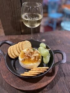 A plate of baked brie cheese with apricot jam and walnuts on top, surrounded by crackers and a small bunch of green grapes. there is a glass of white wine on the table behind the plate of brie.